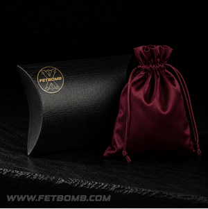 Black pillow-shaped box and red drawstring bag with 'FetBomb' fetish store branding on a dark background.
