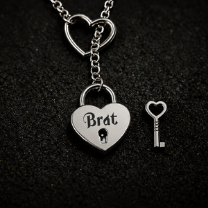 A silver heart-shaped padlock pendant engraved with the word 'Brat' and a small matching heart-shaped key, displayed on a dark textured background.
