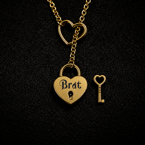 Gold heart-shaped pendant necklace with a lock and key design, featuring the word 'Brat' on the padlock, displayed on a textured black background.