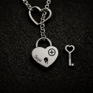A silver heart-shaped lock pendant necklace with a matching heart-shaped key, displayed on a textured black background. The lock is engraved with the name 'Susie' and an owned bdsm female symbol with a heart keyhole symbol.