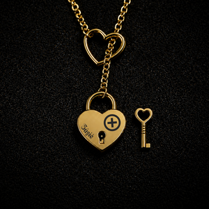 A gold heart-shaped lock pendant necklace on a black textured background, with a matching heart-shaped key beside it. The lock is engraved with the name 'Suzie' and a heart with a plus sign.