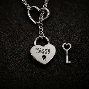 A silver heart-shaped padlock pendant engraved with the name 'Sissy' and a matching heart-shaped key, displayed on a textured black background. BY FETBOMB