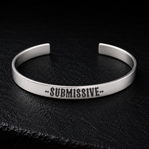 Silver stainless steel cuff bracelet engraved with the word SUBMISSIVE in bold gothic lettering, photographed on a dark textured background, available from FETBOMB.