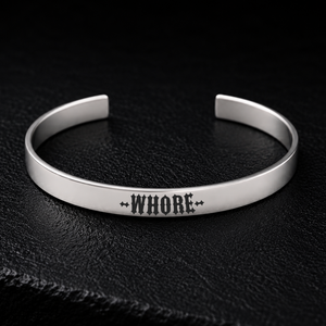 Silver stainless steel cuff bracelet engraved with the word WHORE in bold gothic lettering, photographed on a dark textured background, available from FETBOMB.