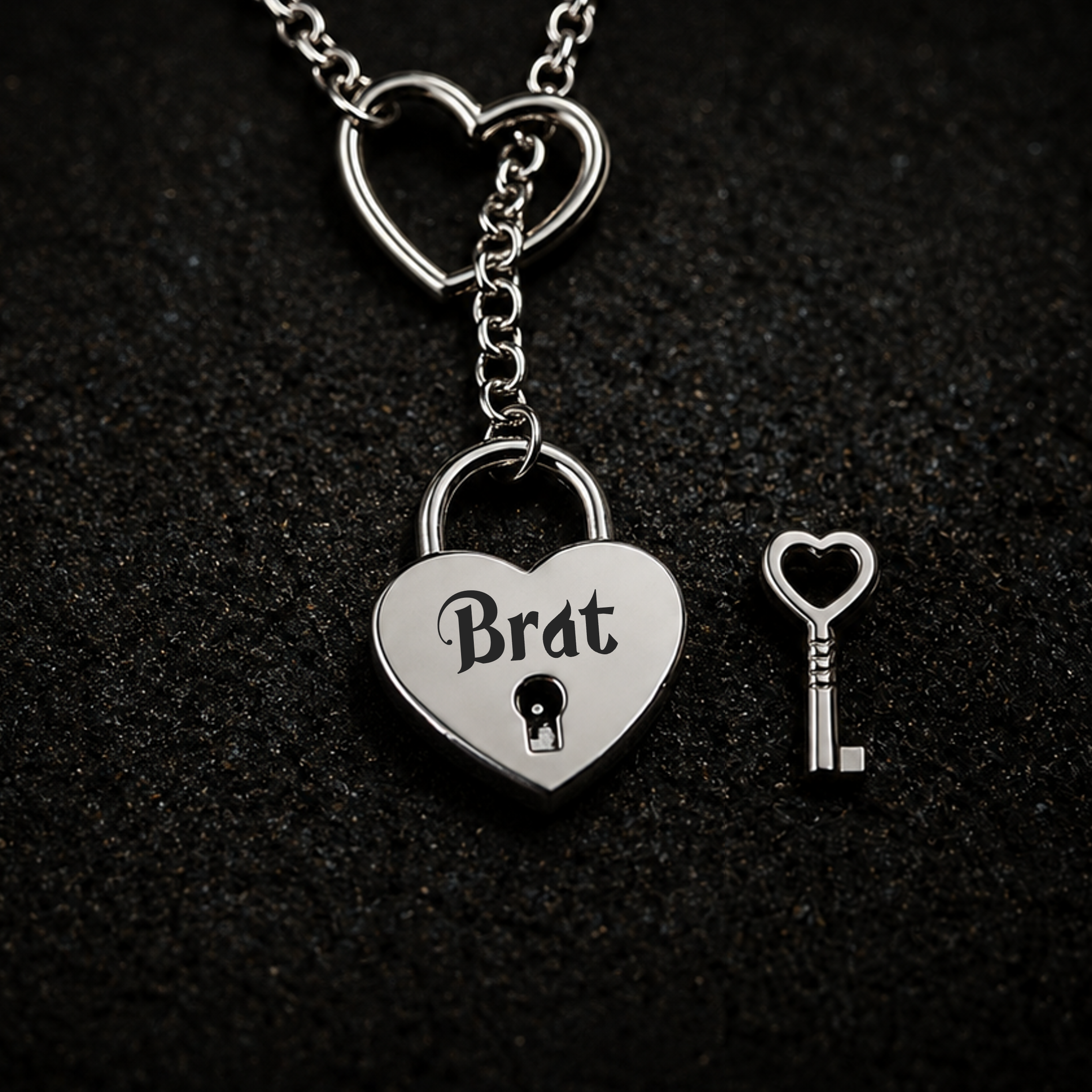 A silver heart-shaped padlock pendant engraved with the word 'Brat' and a small matching heart-shaped key, displayed on a dark textured background.