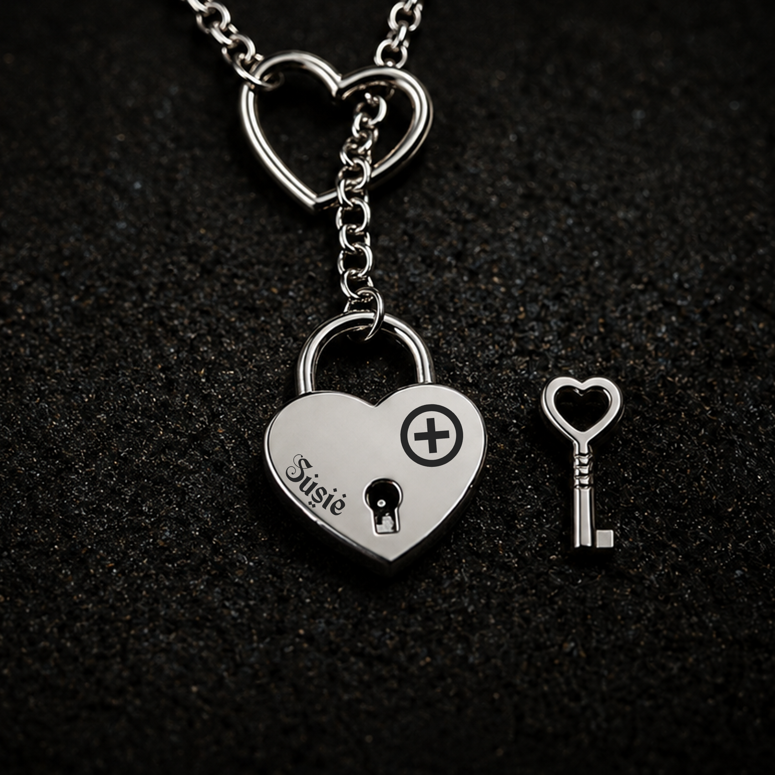 A silver heart-shaped lock pendant necklace with a matching heart-shaped key, displayed on a textured black background. The lock is engraved with the name 'Susie' and an owned bdsm female symbol with a heart keyhole symbol.