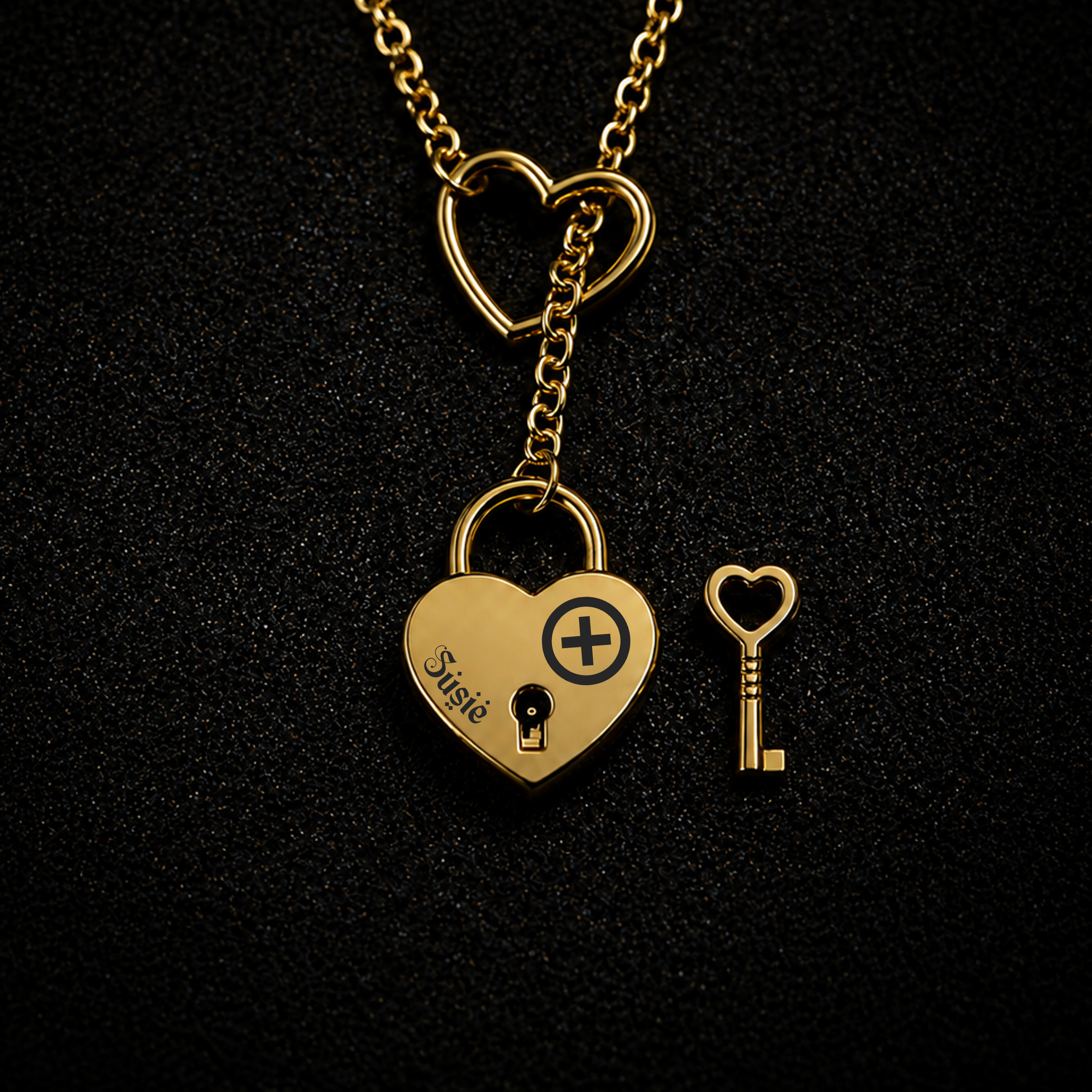 A gold heart-shaped lock pendant necklace on a black textured background, with a matching heart-shaped key beside it. The lock is engraved with the name 'Suzie' and a heart with a plus sign.