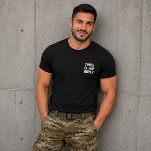 Black personalised owner of pup t-shirt — white bold stacked text "Owner of [pup name]" on left chest, worn by a handler in the pup play community, made to order by FETBOMB.