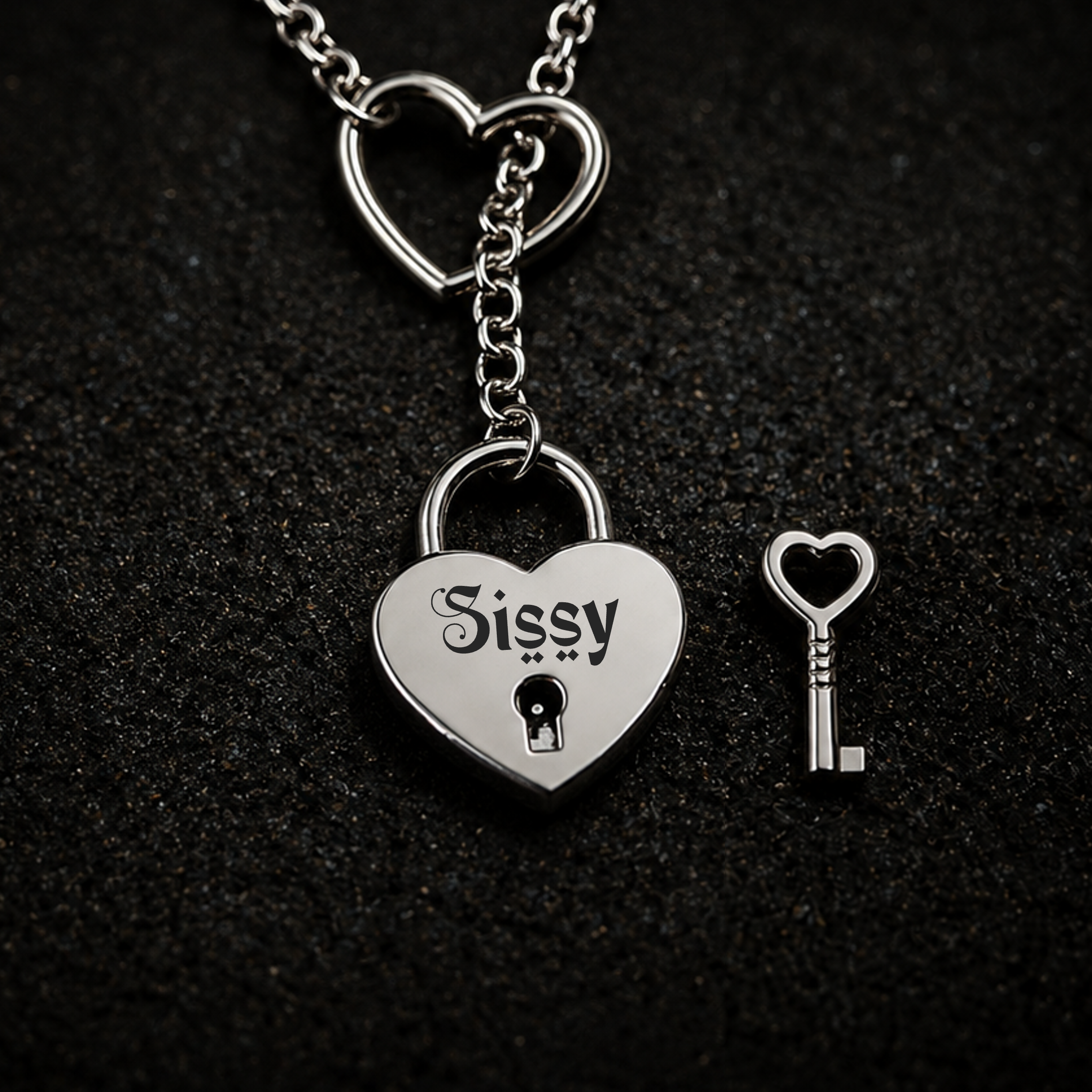 A silver heart-shaped padlock pendant engraved with the name 'Sissy' and a matching heart-shaped key, displayed on a textured black background. BY FETBOMB