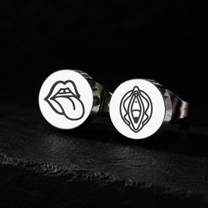 Two stud earrings with a tongue and lips design on one and a logo and vagina on the other, against a black background.