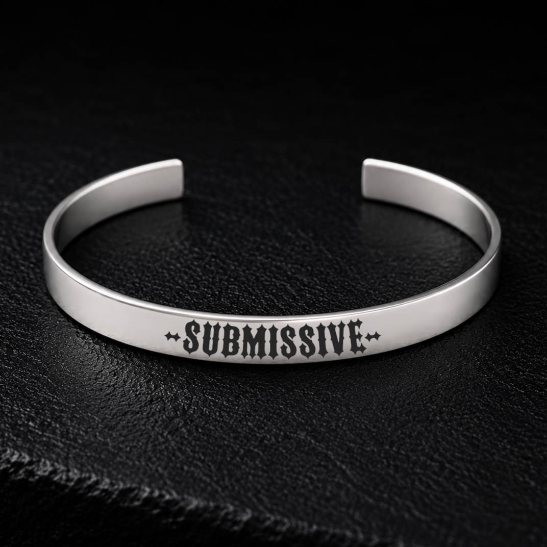 Silver stainless steel cuff bracelet engraved with the word SUBMISSIVE in bold gothic lettering, photographed on a dark textured background, available from FETBOMB.
