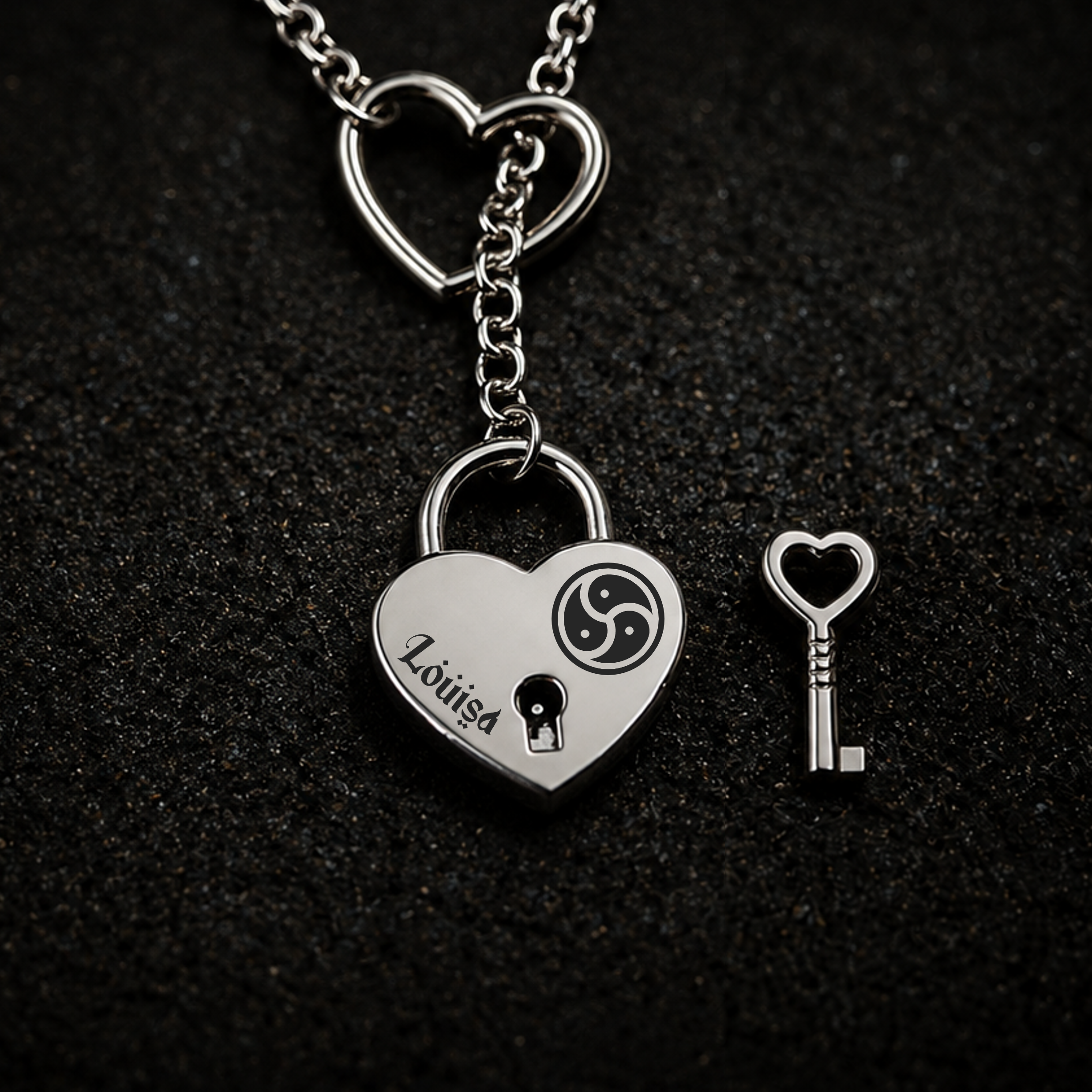 A silver heart-shaped lock pendant necklace with a matching heart-shaped key, displayed on a textured black background. The lock is engraved with a personalised name 'Louisa' and features a black triskelion symbol and keyhole cutout.