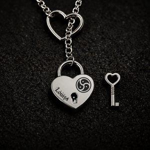 A silver heart-shaped lock pendant necklace with a matching heart-shaped key, displayed on a textured black background. The lock is engraved with a personalised name 'Louisa' and features a black triskelion symbol and keyhole cutout.