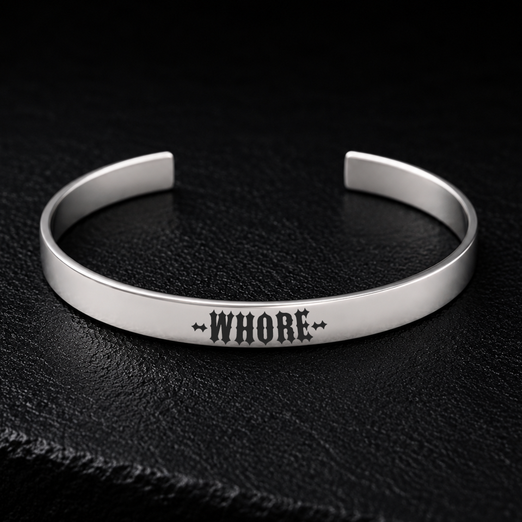Silver stainless steel cuff bracelet engraved with the word WHORE in bold gothic lettering, photographed on a dark textured background, available from FETBOMB.
